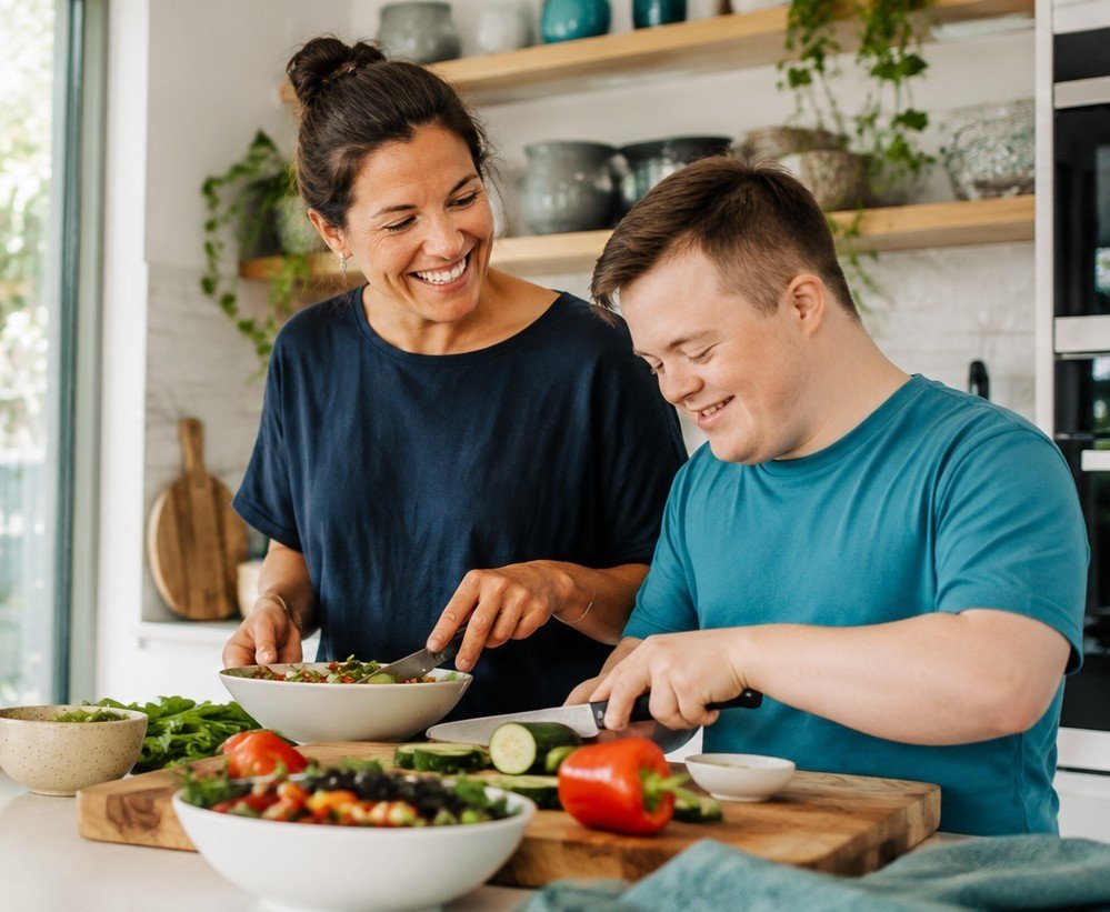 A support worker and a young man laughing together while preparing a meal in a bright home kitchen